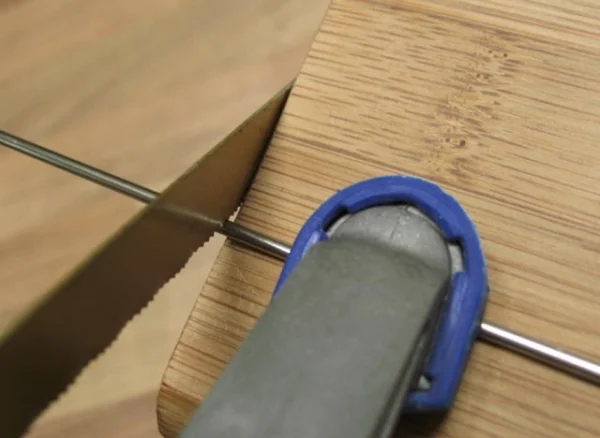 Wellensittich-Bild: Mit der Schraubzwinge fixiert man die Edelstahlstange so auf einem alten Brett oder Ähnlichem, dass man die Metallsäge direkt daran anlegen kann, um nicht abzurutschen. Hätte man diese Führung beim Sägen nicht, entstünden sehr unschöne und rauhe Kratzspuren.. 